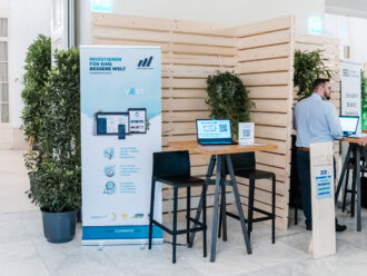 Das Bild zeigt den Messestand von Cleanvest, einer Plattform für nachhaltige Investments. Ein großes, blau-weißes Banner mit dem Slogan „Investieren für eine bessere Welt“ und dem Hashtag #Sauberinvestieren informiert über die Möglichkeit, Fonds nach eigenen Werten zu filtern. Das Banner hebt die Vorteile hervor: hohe Renditechancen, Transparenz und einfache Anwendung. In der Mitte steht ein hoher Stehtisch mit zwei schwarzen Barhockern, auf dem ein Laptop mit geöffneter Cleanvest-Website sowie ein QR-Code zur schnellen Anmeldung platziert sind. Das Holzpaneel im Hintergrund ist mit hübscher grüner Dekoration versehen. Rechts im Bild befindet sich ein weiterer Stand mit der Aufschrift ESG Plus, der sich auf nachhaltige Finanzberatung und Vernetzung konzentriert. Ein Mann in hellblauem Hemd und dunkler Hose steht an einem weiteren Tisch und spricht mit einem Besucher. Der Stand ist mit einer Holzstele mit der Nummer 32 gekennzeichnet.
