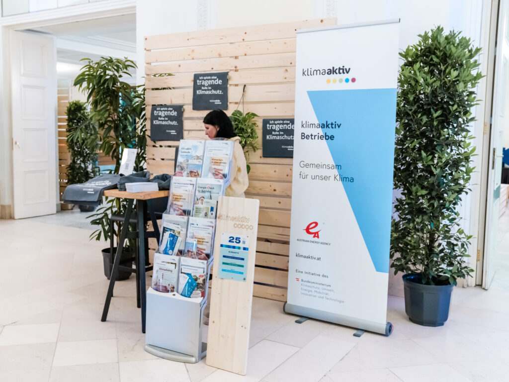 Das Bild zeigt einen Messestand der Initiative klimaaktiv, die sich für Klimaschutz in Unternehmen einsetzt. Ein großes, blau-weißes klimaaktiv-Banner mit dem Slogan „Gemeinsam für unser Klima“ steht rechts neben dem Stand. Eine Frau mit dunklen Haaren, gekleidet in eine hellbeige Jacke, sitzt an einem hohen Stehtisch und scheint sich mit Materialien zu beschäftigen. Auf dem Tisch liegen Flyer, Broschüren und Stofftaschen mit dem klimaaktiv-Logo. Direkt davor befindet sich ein Prospektständer mit zahlreichen Informationsmaterialien zu Themen wie Energieeffizienz, nachhaltige Gebäude und Klimaschutzmaßnahmen. Im Hintergrund sind Holzwände mit Pflanzen dekoriert, und ein schwarzes Schild mit weißem Text trägt die Aufschrift: „Ich spiele eine tragende Rolle im Klimaschutz“. Rechts steht ein hölzernes Schild mit der Standnummer 25.