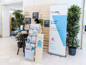 Das Bild zeigt einen Messestand der Initiative klimaaktiv, die sich für Klimaschutz in Unternehmen einsetzt. Ein großes, blau-weißes klimaaktiv-Banner mit dem Slogan „Gemeinsam für unser Klima“ steht rechts neben dem Stand. Eine Frau mit dunklen Haaren, gekleidet in eine hellbeige Jacke, sitzt an einem hohen Stehtisch und scheint sich mit Materialien zu beschäftigen. Auf dem Tisch liegen Flyer, Broschüren und Stofftaschen mit dem klimaaktiv-Logo. Direkt davor befindet sich ein Prospektständer mit zahlreichen Informationsmaterialien zu Themen wie Energieeffizienz, nachhaltige Gebäude und Klimaschutzmaßnahmen. Im Hintergrund sind Holzwände mit Pflanzen dekoriert, und ein schwarzes Schild mit weißem Text trägt die Aufschrift: „Ich spiele eine tragende Rolle im Klimaschutz“. Rechts steht ein hölzernes Schild mit der Standnummer 25.