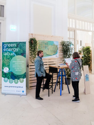 Das Bild zeigt einen Messestand von Green Energy Lab in einer eleganten, lichtdurchfluteten Halle mit hohen Decken. Ein grünes Banner mit dem Slogan „Mit erneuerbaren Energien unseren nicht erneuerbaren Planeten retten!“ und der Webseite www.greenenergylab.at steht links im Vordergrund. Ein Mann mit dunklem Bart und blauem Sakko lehnt entspannt an einem hohen Stehtisch, während er mit einer Frau spricht, die eine auffällig gemusterte Jacke mit roten Details trägt. Sie hält ein Blatt Papier in der Hand und scheint sich über das Thema zu informieren. Im Hintergrund sind Holzwände mit grünen Pflanzen dekoriert, und eine große Landkarte zur Energie-Infrastruktur ist an der Wand befestigt. Ein hölzernes Schild mit der Standnummer 24 steht rechts neben dem Tisch.