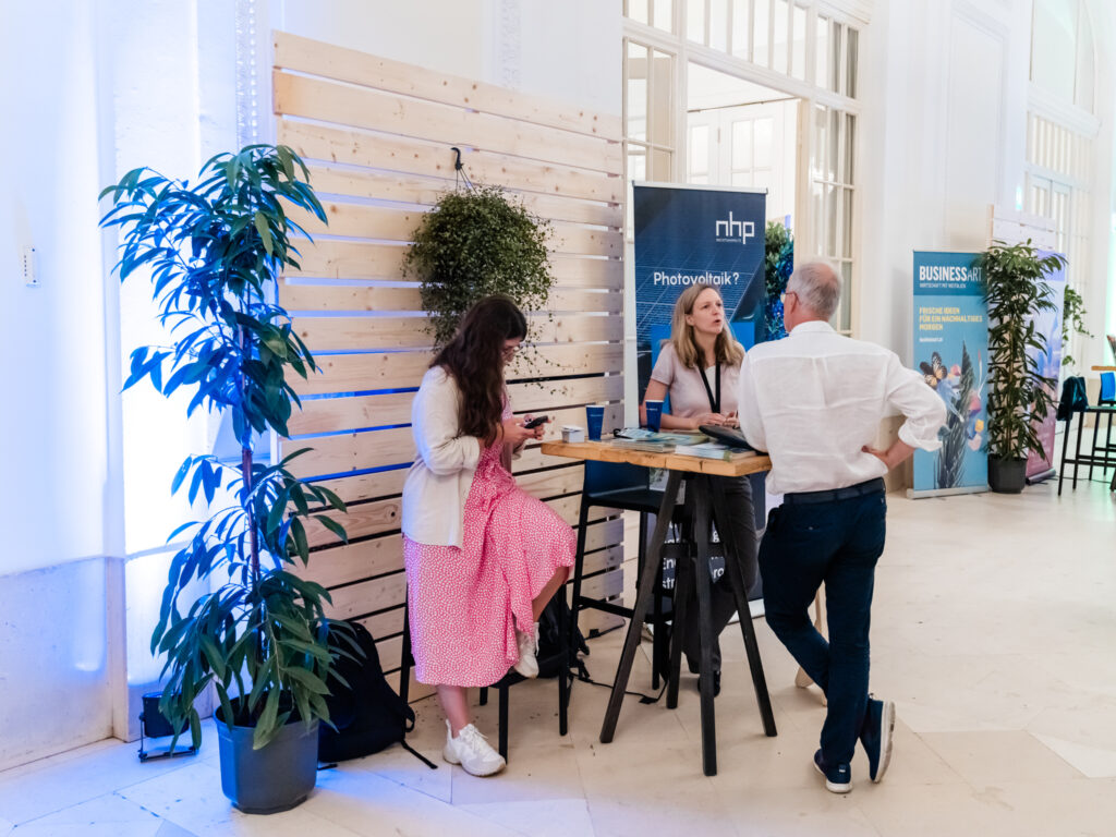 Das Bild zeigt einen Messestand von NHP, der sich mit Photovoltaik beschäftigt. Eine Frau mit langen blonden Haaren, die ein namensschildtragendes Lanyard trägt, steht hinter einem Holztisch und spricht mit einem älteren Mann in weißem Hemd und dunkler Hose, der sich mit einer Hand in die Hüfte gestützt hat. Neben der Szene sitzt eine junge Frau mit langem dunklen Haar, einem rosa-weiß gepunkteten Kleid und weißen Sneakers auf einem Barhocker, während sie auf ihr Smartphone schaut. Im Hintergrund befinden sich weitere Stände, darunter einer mit einem „BUSINESSART“-Banner, der sich mit nachhaltigen Ideen für die Wirtschaft befasst. Die Wände sind mit Holzpaneelen verkleidet, und es gibt große grüne Pflanzen, die mit blauer Beleuchtung in Szene gesetzt sind.