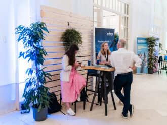 Das Bild zeigt einen Messestand von NHP, der sich mit Photovoltaik beschäftigt. Eine Frau mit langen blonden Haaren, die ein namensschildtragendes Lanyard trägt, steht hinter einem Holztisch und spricht mit einem älteren Mann in weißem Hemd und dunkler Hose, der sich mit einer Hand in die Hüfte gestützt hat. Neben der Szene sitzt eine junge Frau mit langem dunklen Haar, einem rosa-weiß gepunkteten Kleid und weißen Sneakers auf einem Barhocker, während sie auf ihr Smartphone schaut. Im Hintergrund befinden sich weitere Stände, darunter einer mit einem „BUSINESSART“-Banner, der sich mit nachhaltigen Ideen für die Wirtschaft befasst. Die Wände sind mit Holzpaneelen verkleidet, und es gibt große grüne Pflanzen, die mit blauer Beleuchtung in Szene gesetzt sind.