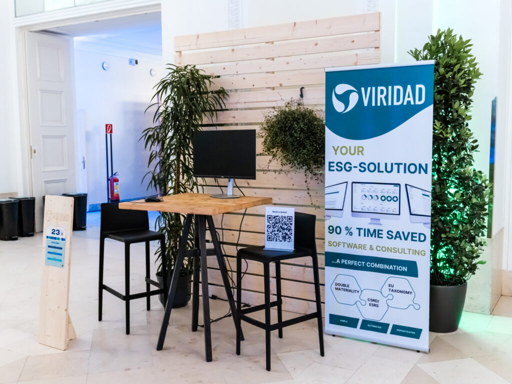 Das Bild zeigt einen Messestand von VIRIDAD, der für seine ESG-Lösung (Environmental, Social, Governance) in Software & Consulting wirbt. Der Banner hebt hervor, dass die Lösung 90 % Zeit spart und sich mit Doppelter Wesentlichkeit, EU-Taxonomie sowie CSRD/ESRS befasst. Die Szene ist modern gestaltet, mit einem Holztisch, zwei schwarzen Barhockern und einem Monitor, der als Präsentationsbildschirm dient. Auf dem Tisch steht ein QR-Code-Schild, das vermutlich weitere Informationen bietet. Die Rückwand besteht aus hellen Holzpaneelen, an denen eine hängende Pflanze befestigt ist, und rechts steht eine große grüne Pflanze, die mit grünem Licht angestrahlt wird. Im Hintergrund ist eine geöffnete Tür zu einem Flur mit blauer Beleuchtung zu sehen. Dort befinden sich mehrere schwarze Abfalleimer zur Mülltrennung sowie ein Feuerlöscher an der Wand. Vor dem Stand steht eine Holztafel mit der Standnummer 23.