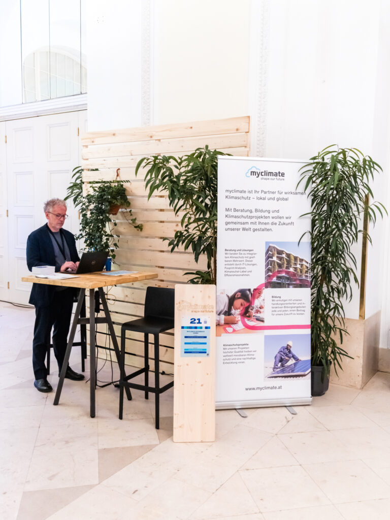 Das Bild zeigt den Messestand von myclimate mit der Standnummer 21 in einem eleganten, hellen Raum mit hohen Decken und weißen Wänden. Ein Mann mit grauen Haaren, dunklem Anzug und Brille steht an einem hohen Stehtisch und arbeitet an seinem Laptop. Neben ihm befindet sich ein großer Roll-up-Banner von myclimate, der die Themen Klimaschutz, Beratung, Bildung und Klimaschutzprojekte präsentiert. Der Banner zeigt Bilder von nachhaltigen Bauprojekten, einem Kind beim Lernen und einem Skifahrer. Im Vordergrund steht eine Holzstele mit der Standnummer 21, und der Bereich ist mit grünen Pflanzen dekoriert. Die Holzwand im Hintergrund sorgt für eine natürliche, umweltbewusste Atmosphäre.
