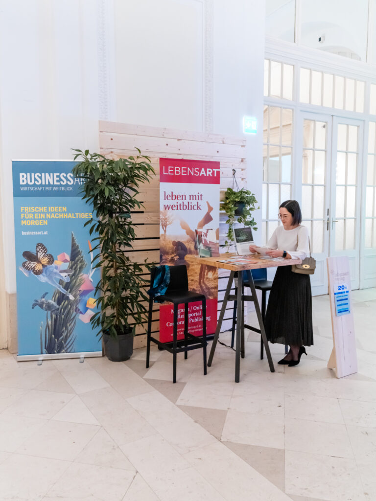 Das Bild zeigt den Messestand von BUSINESSART und LEBENSART mit der Standnummer 20 in einem eleganten, hellen Raum mit großen Fenstern und Türen. Eine Frau mit weißem Oberteil und schwarzem Rock steht an einem hohen Stehtisch, liest ein Magazin und hat eine kleine Umhängetasche dabei. Der Stand besteht aus zwei Roll-up-Bannern: Eines ist blau und trägt die Aufschrift „BUSINESSART – Wirtschaft mit Weitblick“, das andere ist rot und trägt die Aufschrift „LEBENSART – leben mit Weitblick“ mit einem Motiv von Menschen in der Natur. Der Tisch ist mit Magazinen, einem Laptop und einer kleinen Pflanze dekoriert. Daneben steht ein schwarzer Barhocker mit einer blauen Jacke darauf. Im Hintergrund gibt es eine Holzwand mit grünen Pflanzen, die eine natürliche und nachhaltige Atmosphäre schafft.