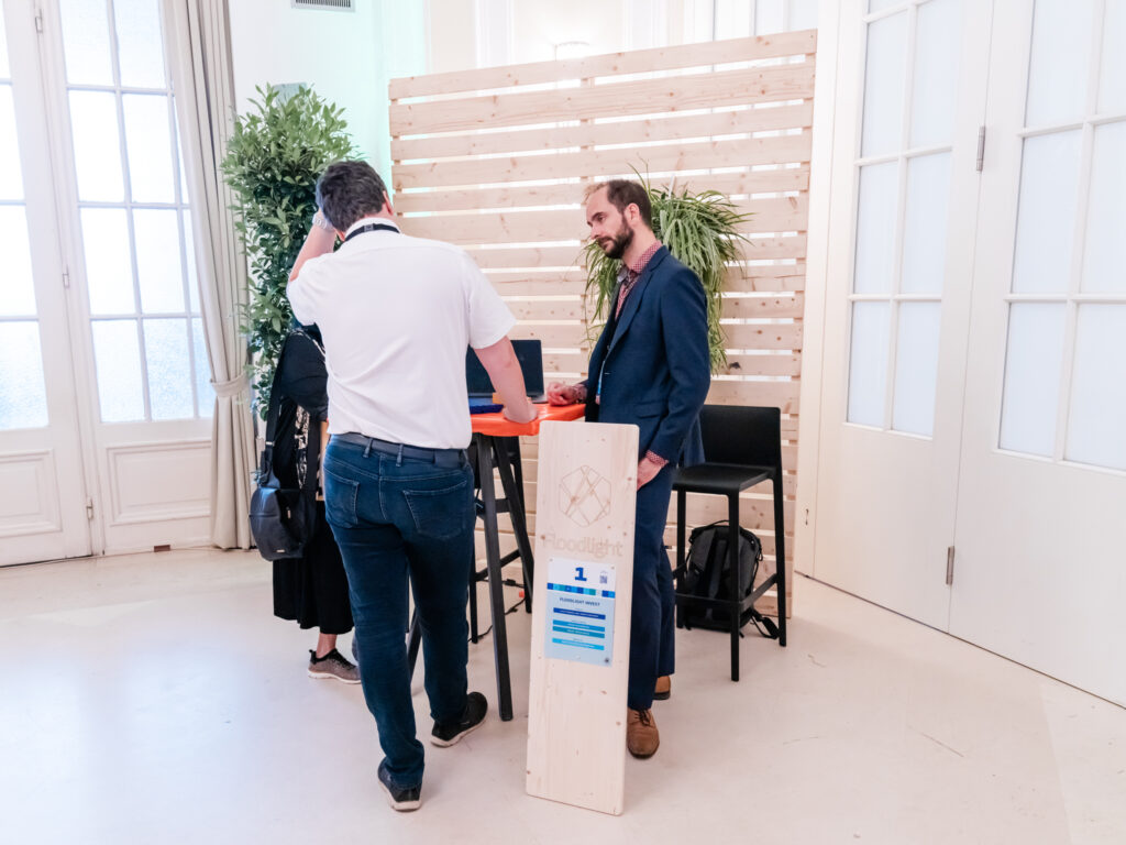 Das Bild zeigt den Messestand von Floodlight Invest mit der Standnummer 1. Ein Mann in einem dunkelblauen Anzug mit rotem Hemd steht hinter einem hohen Stehtisch mit oranger Tischdecke und unterhält sich mit einem Besucher in einem weißen Polohemd und dunkler Jeans. Der Besucher stützt sich mit einer Hand auf den Tisch, während er mit der anderen seinen Kopf berührt. Auf dem Tisch steht ein Laptop, und daneben lehnt ein Holzaufsteller mit dem Floodlight-Logo und den Standinformationen. Im Hintergrund sind Holzwände mit grünen Pflanzen angebracht, die dem Stand eine natürliche und moderne Atmosphäre verleihen. Rechts stehen ein schwarzer Barhocker und eine schwarze Tasche auf dem Boden. Die Szenerie befindet sich in einem hellen, eleganten Raum mit hohen Türen und Fenstern, die für viel Tageslicht sorgen.
