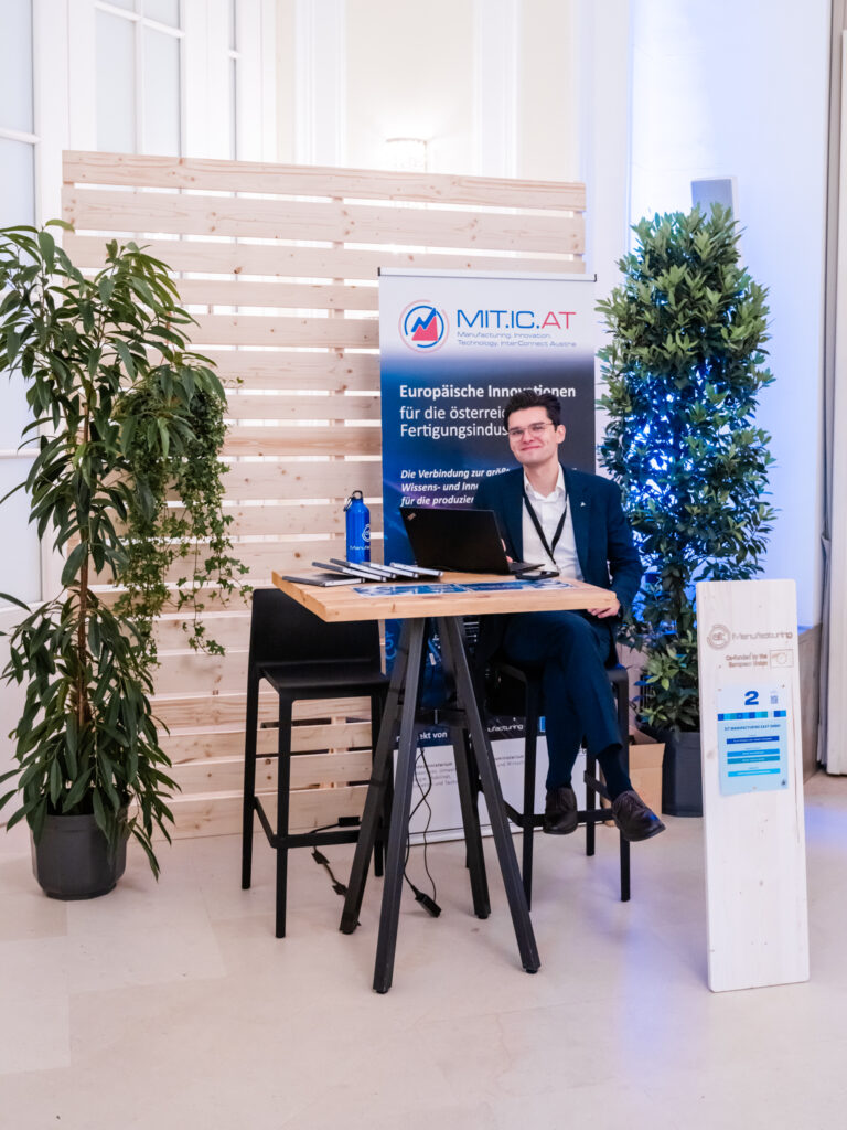 Das Bild zeigt den Messestand von MIT.IC.AT, einer Organisation für europäische Innovationen in der österreichischen Fertigungsindustrie. Ein junger Mann in einem dunklen Anzug und mit Namensschild sitzt entspannt auf einem schwarzen Barhocker, während er freundlich in die Kamera lächelt. Der Stand ist schlicht, aber modern gestaltet, mit einem Banner, das den Namen und die Mission der Organisation präsentiert. Auf dem Holztisch mit schwarzen Metallbeinen befinden sich ein Laptop, einige Dokumente und eine blaue Trinkflasche. Rechts steht ein Holzaufsteller mit der Standnummer „2“, auf dem weitere Informationen zur Organisation zu sehen sind. Um den Stand herum sorgen grüne Pflanzen und Holzwände für eine natürliche und einladende Atmosphäre.