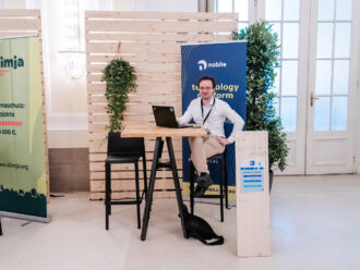 Das Bild zeigt den Messestand der nobile group, einer Technologieplattform, wie auf dem blauen Banner mit goldener Schrift zu lesen ist. Ein Mann in Business-Casual-Kleidung sitzt entspannt an einem hohen Holztisch mit schwarzen Metallbeinen, sein Laptop offen vor sich, während er freundlich in die Kamera lächelt. Der Stand ist schlicht gestaltet, mit einem Holzaufsteller mit der Standnummer „3“, der weitere Informationen über das Unternehmen enthält. Grüne Pflanzen und Holzwände umgeben den Bereich, was eine moderne und nachhaltige Atmosphäre schafft. Links im Bild ist ein weiterer Stand mit einem grünen Banner von „klimja“, das sich mit Klimaschutzprojekten beschäftigt. Die gesamte Szene vermittelt einen professionellen, aber zugänglichen und innovativen Eindruck.