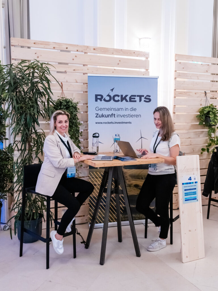 Das Bild zeigt den Messestand von ROCKETS, einem Unternehmen für Investitionen in die Zukunft, wie auf dem Banner zu lesen ist. Zwei Frauen sitzen an einem hohen Holztisch mit schwarzen Metallbeinen, beide mit Laptops vor sich. Sie lächeln und wirken offen für Gespräche mit Messebesuchern. Der Stand ist von Holzpaneelen und grünen Pflanzen umgeben, was eine moderne und nachhaltige Atmosphäre vermittelt. Das Holzschild mit der Standnummer „4“ steht neben dem Tisch und enthält weitere Informationen zum Unternehmen. Das Branding von ROCKETS ist gut sichtbar, mit einem blauen Banner, auf dem eine Windkraftanlage abgebildet ist, was auf eine Ausrichtung auf nachhaltige Investments hindeutet. Die Frauen tragen business-casual Kleidung, was einen professionellen, aber zugänglichen Eindruck hinterlässt.