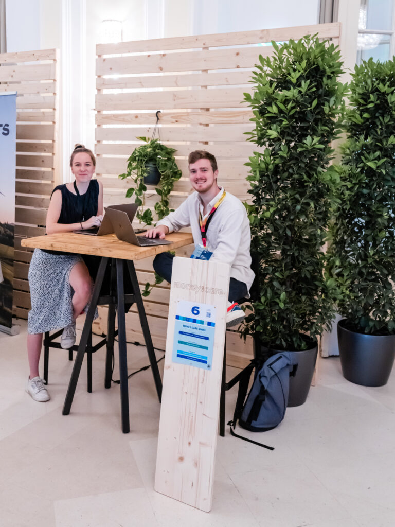 Das Bild zeigt den Messestand von Moneycare, gekennzeichnet durch das Holzschild mit der Standnummer „6“. Zwei junge Personen sitzen an einem hohen Holztisch mit schwarzen Metallbeinen, beide mit Laptops offen. Sie lächeln und wirken freundlich, möglicherweise in einem Gespräch mit Messebesuchern. Der Stand ist von Holzpaneelen und grünen Pflanzen umgeben, was eine natürliche und nachhaltige Atmosphäre schafft. Eine graue Rucksacktasche liegt auf dem Boden neben dem Tisch. Die lockere Kleidung der beiden Personen, darunter Sneakers und ein Sommerkleid, deutet auf eine entspannte, moderne Unternehmensdarstellung hin. Moneycare könnte ein Unternehmen oder eine Initiative im Bereich Finanzen, Nachhaltigkeit oder soziale Verantwortung sein.
