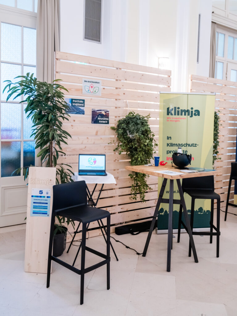Das Bild zeigt den Messestand von Klimja, einer Initiative zur Investition in Klimaschutzprojekte. Der Stand ist mit einem gelb-grünen Banner mit dem Klimja-Logo und dem Slogan „Wir investieren in Morgen“ gestaltet. Auf einem Stehtisch befinden sich Infomaterial und eine schwarze Spendenbox, die wahrscheinlich für eine symbolische Investition oder Spendenaktion genutzt wird. Ein Holzschild mit der Standnummer „5“ steht vor dem Stand. Daneben steht ein kleiner Tisch mit einem Laptop, der eine Grafik zum Thema nachhaltige Finanzen anzeigt. Die Wand ist mit Holzpaneelen und hängenden Pflanzen dekoriert, was eine natürliche und nachhaltige Atmosphäre schafft.
