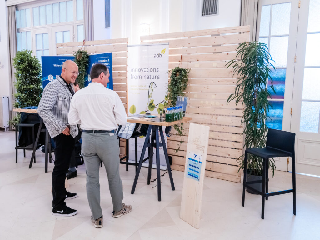 Das Bild zeigt den Messestand des Austrian Centre of Industrial Biotechnology (acib) auf einer Veranstaltung. Der Stand ist mit einem weißen Banner mit grünem Logo und dem Slogan „innovations from nature“ gestaltet. Im Vordergrund stehen zwei Männer im Gespräch; einer trägt ein weißes Hemd und graue Hose, der andere ein gestreiftes Hemd und schwarze Hosen. Auf dem Stehtisch befinden sich mehrere Wasserflaschen, Informationsmaterial und ein kleines Modell einer Pflanze mit Reagenzglas, das biotechnologische Forschung symbolisieren könnte. Links im Bild sind die blauen AWS-Banner eines benachbarten Messestands zu sehen. Eine Holztafel mit der Standnummer „7“ kennzeichnet den acib-Stand. Die gesamte Szene ist von Holzpaneelen mit grünen Pflanzen umgeben, was eine natürliche Atmosphäre schafft.