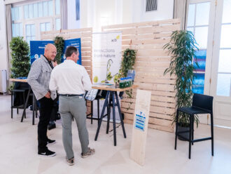 Das Bild zeigt den Messestand des Austrian Centre of Industrial Biotechnology (acib) auf einer Veranstaltung. Der Stand ist mit einem weißen Banner mit grünem Logo und dem Slogan „innovations from nature“ gestaltet. Im Vordergrund stehen zwei Männer im Gespräch; einer trägt ein weißes Hemd und graue Hose, der andere ein gestreiftes Hemd und schwarze Hosen. Auf dem Stehtisch befinden sich mehrere Wasserflaschen, Informationsmaterial und ein kleines Modell einer Pflanze mit Reagenzglas, das biotechnologische Forschung symbolisieren könnte. Links im Bild sind die blauen AWS-Banner eines benachbarten Messestands zu sehen. Eine Holztafel mit der Standnummer „7“ kennzeichnet den acib-Stand. Die gesamte Szene ist von Holzpaneelen mit grünen Pflanzen umgeben, was eine natürliche Atmosphäre schafft.