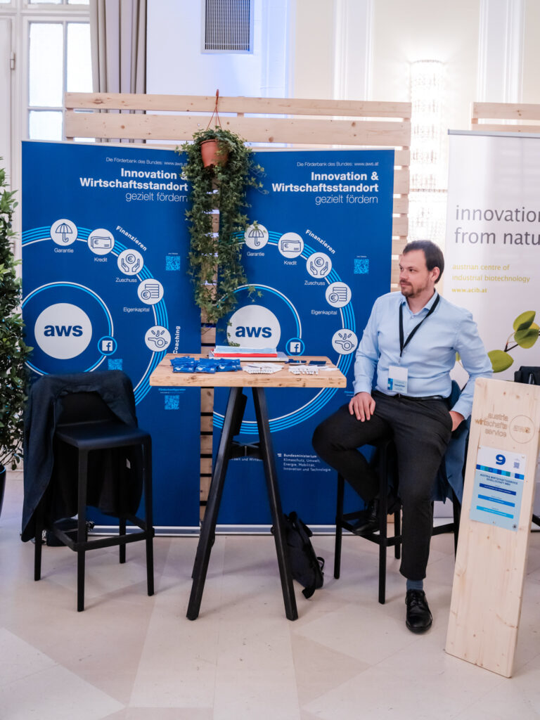 Das Bild zeigt den AWS-Messestand auf einer Veranstaltung zur Innovations- und Wirtschaftsstandortförderung. Der Stand besteht aus zwei blauen Bannern mit dem AWS-Logo und Symbolen für Finanzierungsangebote wie Garantien, Kredite, Zuschüsse und Eigenkapital. Zwischen den Bannern hängt eine grüne Pflanze. Ein Mann in hellblauem Hemd und dunkler Hose sitzt entspannt auf einem Barhocker am Stehtisch, trägt ein Namensschild und scheint als Ansprechpartner am Stand zu sein. Der Tisch ist mit Flyern, Stiften und Werbeartikeln bestückt. Links neben ihm steht ein weiterer leerer Barhocker mit einer Jacke darüber. Neben dem AWS-Stand ist ein weiteres Banner des Austrian Centre of Industrial Biotechnology (ACIB) sichtbar, das auf nachhaltige Innovationen aus der Natur hinweist. Eine Holztafel mit der Standnummer „9“ markiert den AWS-Stand.