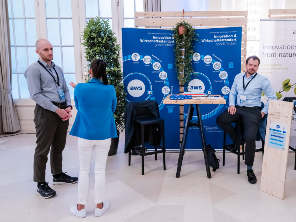 Das Bild zeigt einen Messestand von AWS (Austria Wirtschaftsservice) auf einer Veranstaltung. Der Stand ist mit einem großen blauen Banner gestaltet, das die Fördermöglichkeiten für Innovation und den Wirtschaftsstandort erklärt. Darauf sind Icons für Finanzierungen, Garantien, Kredite und Eigenkapital abgebildet. Zwei Männer mit Namensschildern und eine Frau im blauen Blazer und weißen Hosen stehen im Gespräch. Einer der Männer sitzt auf einem Barhocker, während der andere steht. Der Stand ist mit einem Holzhintergrund, Pflanzen und einem Stehtisch gestaltet. Daneben befindet sich eine Holztafel mit der Standnummer „9“, die AWS als Aussteller markiert. Im Hintergrund ist ein weiteres Banner von ACIB (Austrian Centre of Industrial Biotechnology) zu sehen.