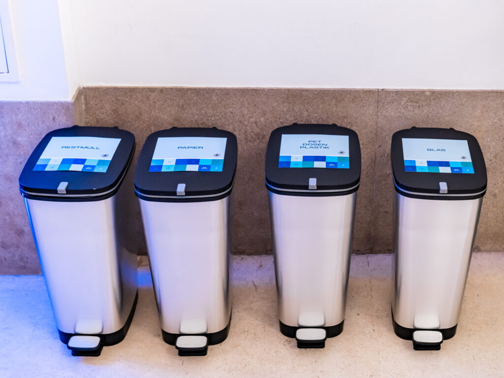 Das Bild zeigt vier moderne Mülleimer aus Edelstahl mit schwarzen Deckeln, die jeweils mit einem digitalen Display versehen sind. Die Displays zeigen die Abfallkategorien: Restmüll Papier PET, Dosen, Plastik Glas Jeder Mülleimer hat ein Fußpedal zum Öffnen. Die Mülleimer stehen an einer beigen Steinwand in einem hell beleuchteten Raum. Das Licht wirft einen leichten blauen Schimmer auf den Boden, was auf eine moderne oder nachhaltige Veranstaltung hinweisen könnte.