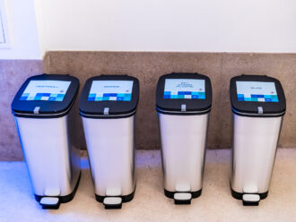 Das Bild zeigt vier moderne Mülleimer aus Edelstahl mit schwarzen Deckeln, die jeweils mit einem digitalen Display versehen sind. Die Displays zeigen die Abfallkategorien: Restmüll Papier PET, Dosen, Plastik Glas Jeder Mülleimer hat ein Fußpedal zum Öffnen. Die Mülleimer stehen an einer beigen Steinwand in einem hell beleuchteten Raum. Das Licht wirft einen leichten blauen Schimmer auf den Boden, was auf eine moderne oder nachhaltige Veranstaltung hinweisen könnte.