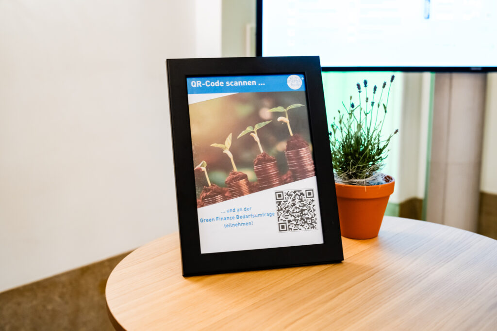 Das Bild zeigt einen schwarzen Bilderrahmen mit einem Poster, das zur Teilnahme an einer Green Finance Bedarfsumfrage einlädt. Der obere Teil des Posters trägt die Aufschrift „QR-Code scannen …“ und enthält das Logo des Klima- und Energiefonds. Das zentrale Bild auf dem Poster zeigt gestapelte Münzen, auf denen kleine grüne Pflanzen keimen, was Wachstum und nachhaltige Investitionen symbolisiert. Unten befindet sich ein QR-Code, mit dem die Umfrage aufgerufen werden kann. Neben dem Rahmen steht ein kleiner Lavendeltopf in einem orangen Blumentopf auf einem runden Holztisch. Im Hintergrund ist ein Bildschirm mit verschwommener Anzeige zu sehen.