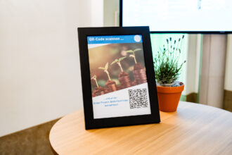Das Bild zeigt einen schwarzen Bilderrahmen mit einem Poster, das zur Teilnahme an einer Green Finance Bedarfsumfrage einlädt. Der obere Teil des Posters trägt die Aufschrift „QR-Code scannen …“ und enthält das Logo des Klima- und Energiefonds. Das zentrale Bild auf dem Poster zeigt gestapelte Münzen, auf denen kleine grüne Pflanzen keimen, was Wachstum und nachhaltige Investitionen symbolisiert. Unten befindet sich ein QR-Code, mit dem die Umfrage aufgerufen werden kann. Neben dem Rahmen steht ein kleiner Lavendeltopf in einem orangen Blumentopf auf einem runden Holztisch. Im Hintergrund ist ein Bildschirm mit verschwommener Anzeige zu sehen.
