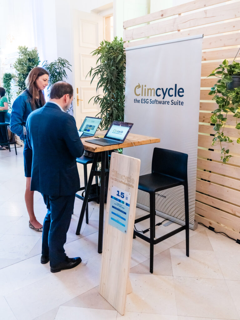 Das Bild zeigt den Messestand von Climcycle, einer ESG-Software-Suite. Ein großes Banner mit dem Logo von Climcycle steht hinter einem hohen Holztisch, an dem zwei Laptops geöffnet sind. Ein Mann in einem dunklen Anzug betrachtet den Bildschirm, während eine Frau in einem dunkelblauen Outfit danebensteht. Der Stand trägt die Nummer 15, die auf einem Holzschild vor dem Tisch zu sehen ist. Die Umgebung ist mit Holzverkleidung und Pflanzen gestaltet, was eine nachhaltige Atmosphäre vermittelt.