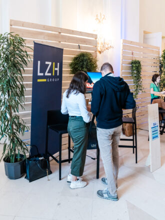 Das Bild zeigt einen Messestand der LZH Group mit der Standnummer 11.1. Zwei Personen stehen vor einem Monitor, der auf einem hohen schwarzen Tisch platziert ist. Die Frau trägt eine weiße Bluse und grüne Hose, während der Mann einen schwarzen Hoodie und beige Hose trägt. Der Stand ist mit einer Holzwand und grünen Pflanzen dekoriert, was eine nachhaltige und moderne Atmosphäre schafft. Links neben dem Tisch steht eine große schwarze Tasche mit einem Schlüsselanhänger. Im Hintergrund ist ein weiterer Stand mit einer Frau, die an einem Laptop arbeitet.
