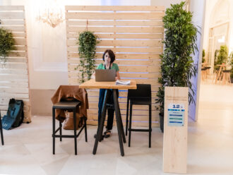 Das Bild zeigt einen Messestand von Grünes Gold mit der Standnummer 12. Eine Frau mit kurzen dunklen Haaren sitzt an einem hohen Holztisch mit einem Laptop und einigen Unterlagen. Der Stand ist nachhaltig gestaltet, mit Holzwänden und viel grüner Bepflanzung. Neben dem Tisch stehen zwei hohe schwarze Hocker, auf einem liegt eine braune Jacke. Im Hintergrund sind weitere Stände und Pflanzen zu sehen, die eine offene und einladende Atmosphäre schaffen.