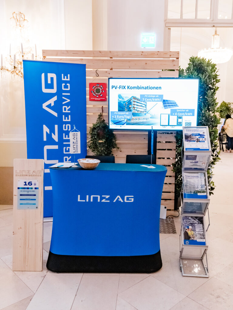 Das Bild zeigt einen Messestand der LINZ AG Energieservice in einer eleganten Halle mit hohen Decken. Der Stand ist in Blau gehalten und umfasst einen Empfangstisch mit dem LINZ AG Logo, eine große Werbetafel mit Informationen zu PV-FIX Kombinationen für Photovoltaik-Tarife sowie einen Bildschirm mit weiteren Details. Ein Prospektständer mit Infomaterialien steht auf der rechten Seite. Eine Holztafel mit der Nummer 16 deutet auf einen Standplatz innerhalb einer größeren Veranstaltung hin. Begrünungselemente und eine hölzerne Rückwand verleihen dem Stand eine moderne, nachhaltige Ästhetik.
