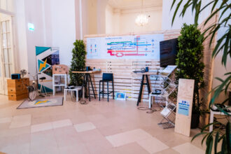 Das Bild zeigt eine modern gestaltete Ausstellung oder einen Informationsstand zu erneuerbaren Energien in einem eleganten, hellen Raum mit hohen Decken und Kronleuchter. Der Stand besteht aus einer Holzwand mit einem großen Infoposter zum Thema „Energiefluss in Österreich 2022“, daneben ein Bildschirm sowie verschiedene interaktive Elemente. Links steht ein Ergometer-Fahrrad, das vermutlich zur Demonstration von Energieerzeugung genutzt wird. Pflanzen und Holzmaterialien schaffen eine nachhaltige Atmosphäre, während Stühle und Tische zur Interaktion einladen. Der Stand trägt den Namen „Power Playground“.