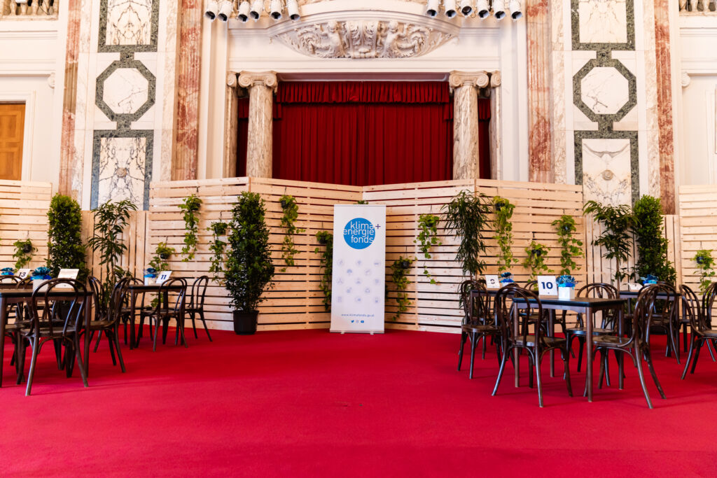 Das Bild zeigt einen eleganten Veranstaltungsbereich mit einem roten Teppich, umgeben von klassischen Marmorwänden mit goldenen und grünen Verzierungen. Im Zentrum befindet sich eine Bühne mit einem roten Vorhang, eingerahmt von zwei Marmorsäulen. Davor stehen mehrere dunkle Holzstühle und Tische, die mit blauen Blumen in weißen Töpfen dekoriert sind. An den Tischen sind Nummern wie 