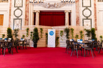 Das Bild zeigt einen eleganten Veranstaltungsbereich mit einem roten Teppich, umgeben von klassischen Marmorwänden mit goldenen und grünen Verzierungen. Im Zentrum befindet sich eine Bühne mit einem roten Vorhang, eingerahmt von zwei Marmorsäulen. Davor stehen mehrere dunkle Holzstühle und Tische, die mit blauen Blumen in weißen Töpfen dekoriert sind. An den Tischen sind Nummern wie 