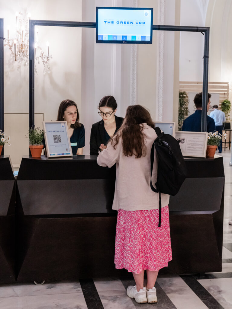 Eine Besucherin mit einem schwarzen Rucksack und einem pink gemusterten Rock steht an einem Empfangsschalter mit der Aufschrift 'THE GREEN 100'. Zwei Mitarbeiterinnen hinter dem Schalter helfen ihr bei der Registrierung. Auf dem Schalter befinden sich kleine Topfpflanzen und Informationsmaterialien. Ein Bildschirm über dem Schalter zeigt ebenfalls das 'THE GREEN 100'-Logo. Im Hintergrund sind ein Kronleuchter und weitere Teilnehmer zu erkennen, die sich im Empfangsbereich bewegen.