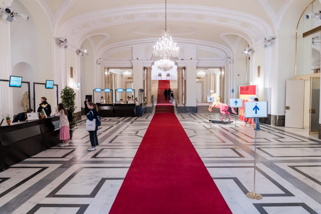 Eine elegante Empfangshalle mit einem langen roten Teppich, der durch die Mitte des Raums zu einer breiten Treppe mit roten Stufen führt. Links befinden sich mehrere Check-in-Schalter mit Bildschirmen, auf denen 'THE GREEN 100' angezeigt wird. Einige Besucherinnen stehen an den Schaltern, um sich zu registrieren. Rechts sind Richtungsschilder mit blauen Pfeilen sowie eine auffällige, pinkfarbene Pferdeskulptur zu sehen. Der Raum ist mit Marmorboden, großen Spiegeln, hohen Decken und prunkvollen Kronleuchtern geschmückt, was eine edle Atmosphäre schafft.