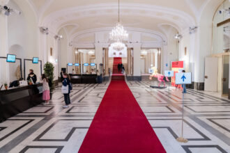 Eine elegante Empfangshalle mit einem langen roten Teppich, der durch die Mitte des Raums zu einer breiten Treppe mit roten Stufen führt. Links befinden sich mehrere Check-in-Schalter mit Bildschirmen, auf denen 'THE GREEN 100' angezeigt wird. Einige Besucherinnen stehen an den Schaltern, um sich zu registrieren. Rechts sind Richtungsschilder mit blauen Pfeilen sowie eine auffällige, pinkfarbene Pferdeskulptur zu sehen. Der Raum ist mit Marmorboden, großen Spiegeln, hohen Decken und prunkvollen Kronleuchtern geschmückt, was eine edle Atmosphäre schafft.