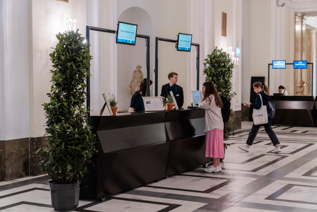 Eine Empfangshalle mit mehreren Informationsschaltern, an denen Bildschirme mit der Aufschrift 'THE GREEN 100' angebracht sind. Zwei Mitarbeitende, ein Mann und eine Frau, helfen Besuchern am Schalter, während eine junge Frau in pinkem Rock und grauer Jacke mit Rucksack sich nach vorne lehnt, um Informationen zu erhalten. Im Hintergrund geht eine weitere Frau mit einer 'our power'-Tasche vorbei. Die Umgebung ist hell mit Marmorfußboden, hohen weißen Wänden und eleganten Pflanzen, die den Raum optisch auflockern.
