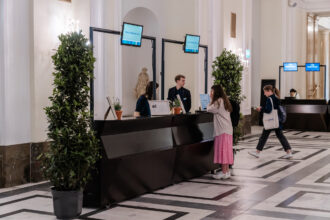 Eine Empfangshalle mit mehreren Informationsschaltern, an denen Bildschirme mit der Aufschrift 'THE GREEN 100' angebracht sind. Zwei Mitarbeitende, ein Mann und eine Frau, helfen Besuchern am Schalter, während eine junge Frau in pinkem Rock und grauer Jacke mit Rucksack sich nach vorne lehnt, um Informationen zu erhalten. Im Hintergrund geht eine weitere Frau mit einer 'our power'-Tasche vorbei. Die Umgebung ist hell mit Marmorfußboden, hohen weißen Wänden und eleganten Pflanzen, die den Raum optisch auflockern.