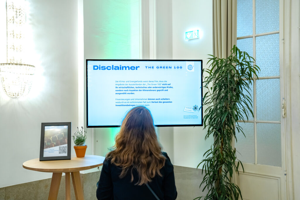 Eine Frau mit langen, braunen Haaren steht vor einem großen Bildschirm und liest einen Haftungsausschluss mit der Überschrift 'Disclaimer' im Rahmen der Veranstaltung 'The Green 100'. Der Text weist darauf hin, dass die Angebote der Aussteller nicht nach wirtschaftlichen oder technischen Risiken, sondern nach Klimarelevanz ausgewählt wurden. Neben dem Bildschirm befindet sich ein runder Holztisch mit einem kleinen Blumentopf und einem gerahmten Informationsschild, das einen QR-Code zeigt. Im Hintergrund sind eine große grüne Pflanze und ein Notausgangsschild sichtbar, die dem Raum eine freundliche Atmosphäre verleihen.