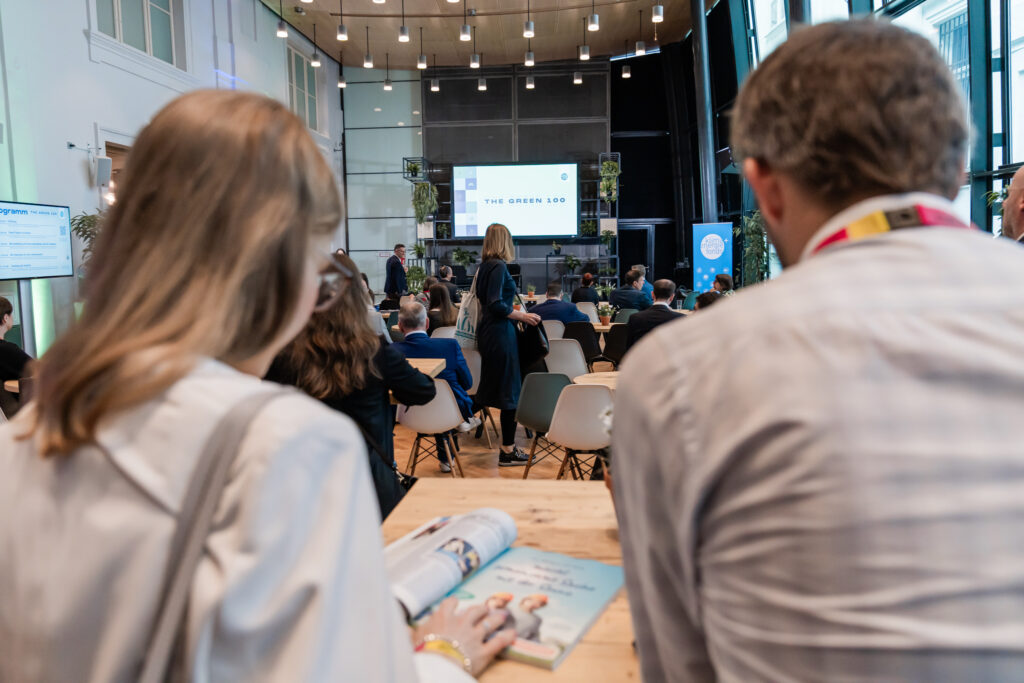 Blick von hinten in einen Konferenzraum, in dem zahlreiche Personen an runden Tischen sitzen und auf eine Bühne mit einer großen Leinwand blicken, auf der 'THE GREEN 100' angezeigt wird. Im Publikum befinden sich Menschen in Businesskleidung, einige stehen und unterhalten sich. Im Vordergrund sind zwei Personen von hinten zu sehen, eine davon hält ein geöffnetes Magazin in der Hand. Der Veranstaltungsort ist modern gestaltet mit großen Fenstern, hängenden Lampen und vielen Grünpflanzen.