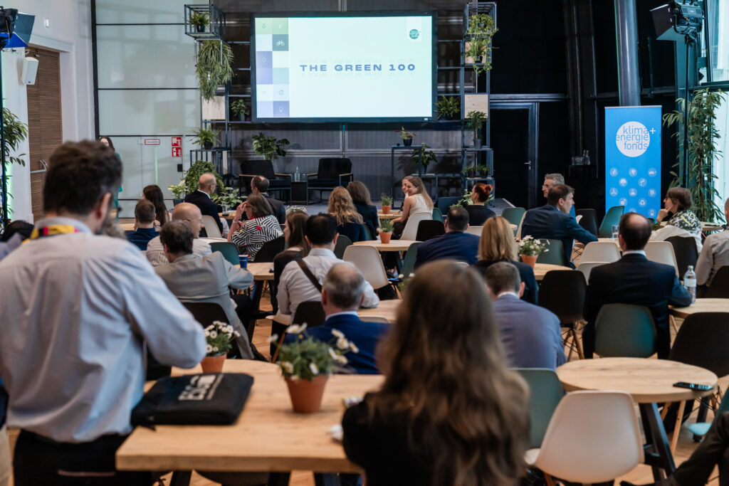 Ein Konferenzraum mit mehreren runden Holztischen, an denen Menschen sitzen und einer Präsentation folgen. Im Hintergrund ist eine große Leinwand zu sehen, auf der 'THE GREEN 100' steht. Der Raum ist modern gestaltet, mit vielen grünen Pflanzen als Dekoration. Auf der rechten Seite steht ein Roll-up mit dem Logo von 'Klima+Energiefonds'.