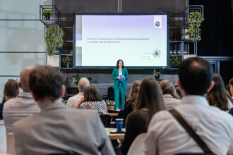 Eine Frau in einem smaragdgrünen Hosenanzug steht auf einer Bühne vor einem Publikum und hält eine Präsentation. Sie hält ein blaues Programmheft und trägt ein Headset-Mikrofon. Hinter ihr zeigt eine große Leinwand den Text: 'Die Veranstaltung entspricht den Kriterien des Österreichischen Umweltzeichens für Green Events.' Der Raum ist modern gestaltet, mit vielen grünen Pflanzen als Dekoration.