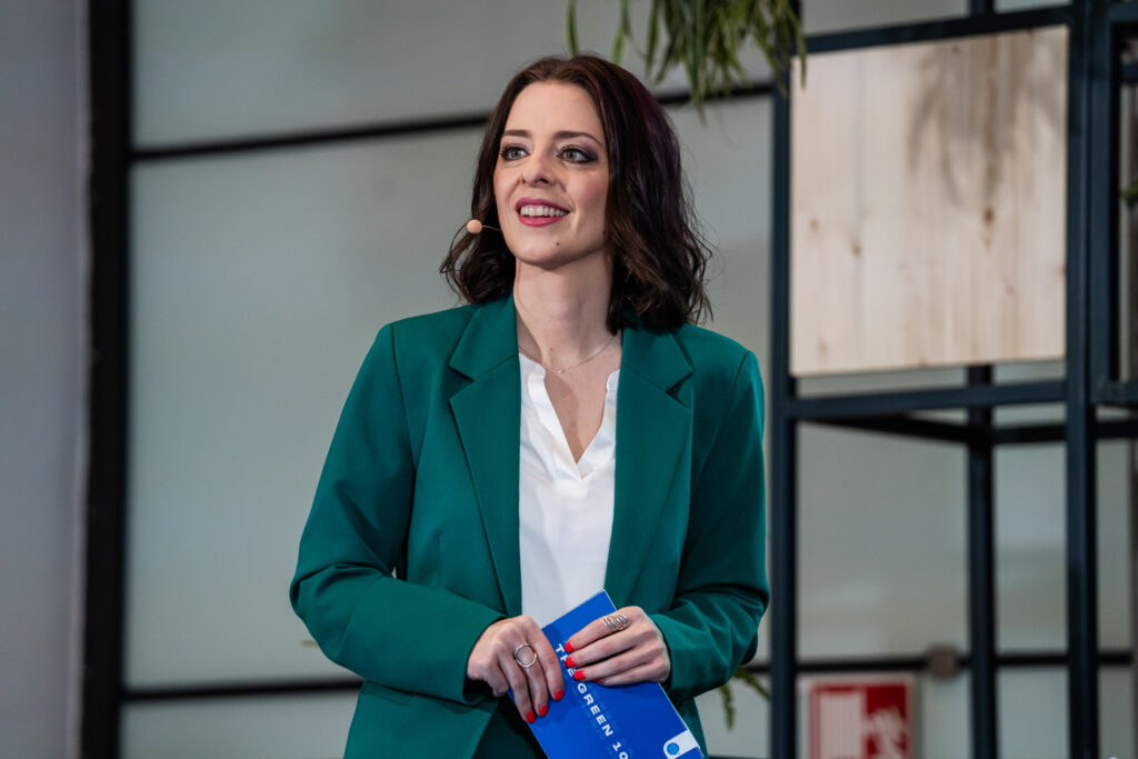 Eine Frau mit schulterlangem braunem Haar steht auf einer Bühne. Sie trägt ein smaragdgrünes Sakko über einer weißen Bluse und hält ein blaues Programmheft mit der Aufschrift 'THE GREEN'. Sie trägt ein Headset-Mikrofon und lächelt, während sie nach vorne blickt. Im Hintergrund sind unscharf moderne Regale mit Holz- und Pflanzendekoration zu sehen.