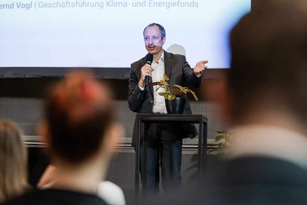 Ein Mann in einem dunklen Anzug mit weißem Hemd hält ein Mikrofon in der rechten Hand und spricht auf einer Bühne. Er steht neben einem kleinen Tisch, auf dem ein Blumentopf mit einer Pflanze steht. Im Hintergrund sind weitere grüne Pflanzen sowie ein Regal mit Holzpaneelen zu sehen. Der Mann lächelt leicht und scheint in eine Präsentation oder Rede vertieft zu sein.