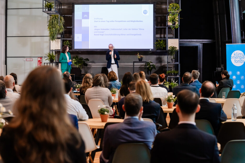 Eine Konferenzszene mit einem Mann und einer Frau auf der Bühne. Der Mann trägt ein dunkelblaues Sakko, ein weißes Hemd und helle Hosen, während die Frau in einem grünen Hosenanzug gekleidet ist. Beide halten Mikrofone und sprechen vor einem Publikum. Im Hintergrund ist eine große Leinwand mit der Überschrift 'Eröffnung: Start in einen Tag voller Perspektiven und Möglichkeiten' zu sehen. Das Publikum sitzt an Tischen mit kleinen Blumentöpfen, und die Umgebung ist mit Pflanzen dekoriert. Auf der rechten Seite steht ein blaues Banner mit der Aufschrift 'Klima + Energiefonds'.