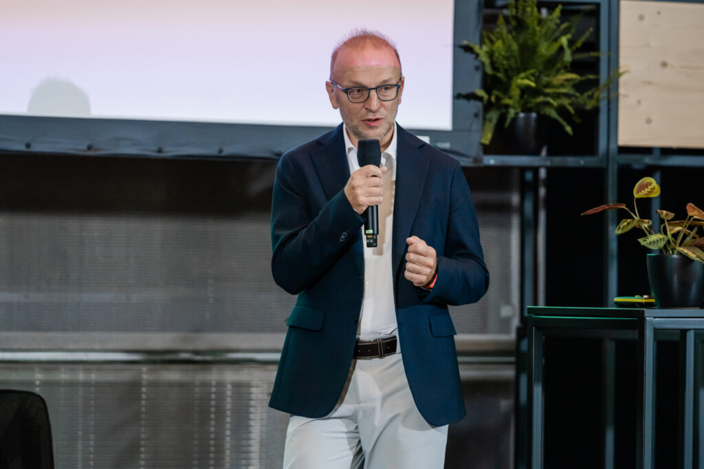 Ein Mann in einem dunkelblauen Sakko, weißem Hemd und hellen Hosen hält ein Mikrofon und spricht auf einer Bühne. Er trägt eine Brille und eine rote Armbanduhr. Im Hintergrund befinden sich einige grüne Pflanzen und ein großes Display, das teilweise zu sehen ist. Die Szene wirkt professionell, als ob er eine Präsentation oder Rede bei einer Konferenz hält.
