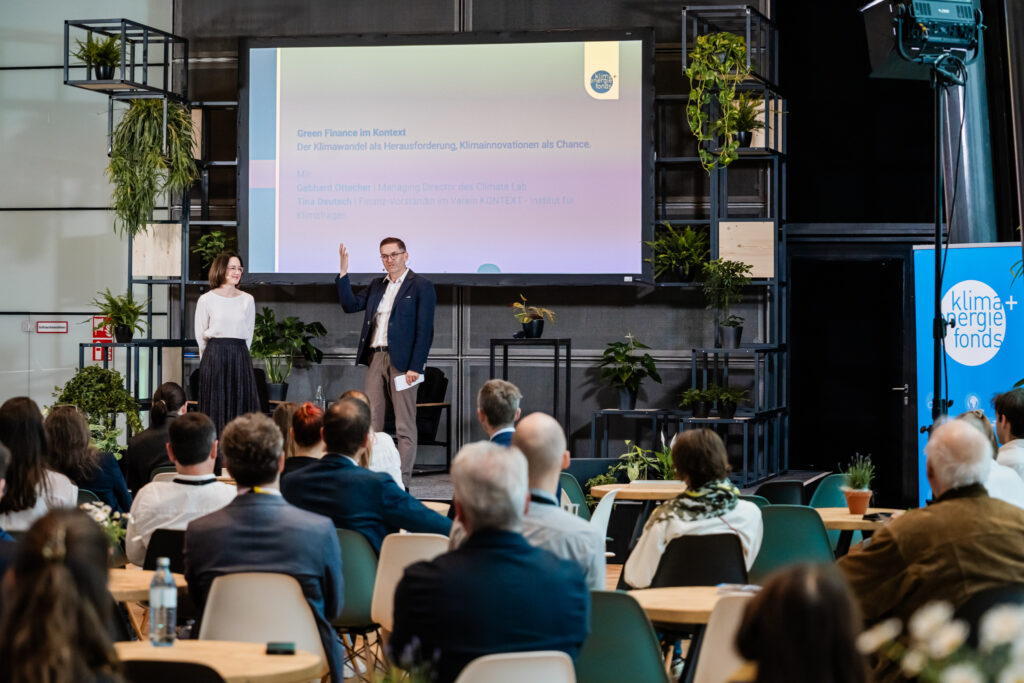 Ein Mann und eine Frau stehen auf einer Bühne vor einem Publikum. Im Hintergrund ist eine große Präsentationsfolie mit dem Titel 'Green Finance im Kontext: Der Klimawandel als Herausforderung, Klimainnovationen als Chance' zu sehen. Der Mann trägt ein dunkelblaues Sakko und hebt eine Hand, während die Frau in einer weißen Bluse und einem dunklen Rock lächelt. Der Raum ist mit grünen Pflanzen dekoriert, und im Vordergrund sitzen zahlreiche Zuhörer. Rechts im Bild steht ein Banner mit der Aufschrift 'Klima + Energie Fonds'.