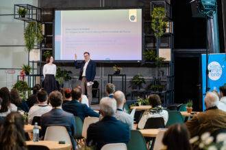 Ein Mann und eine Frau stehen auf einer Bühne vor einem Publikum. Im Hintergrund ist eine große Präsentationsfolie mit dem Titel 'Green Finance im Kontext: Der Klimawandel als Herausforderung, Klimainnovationen als Chance' zu sehen. Der Mann trägt ein dunkelblaues Sakko und hebt eine Hand, während die Frau in einer weißen Bluse und einem dunklen Rock lächelt. Der Raum ist mit grünen Pflanzen dekoriert, und im Vordergrund sitzen zahlreiche Zuhörer. Rechts im Bild steht ein Banner mit der Aufschrift 'Klima + Energie Fonds'.