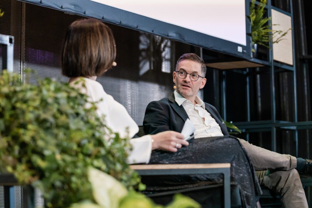 Ein Mann und eine Frau sitzen auf einer Bühne in einer Gesprächsrunde. Beide tragen Headset-Mikrofone. Der Mann mit Brille, dunklem Sakko und weißem Hemd schaut aufmerksam zur Frau, die von hinten zu sehen ist. Im Vordergrund verdecken grüne Pflanzen teilweise die Sicht. Im Hintergrund sind Metallregale mit weiteren Pflanzen sichtbar, was der Szene eine moderne, offene Atmosphäre verleiht.