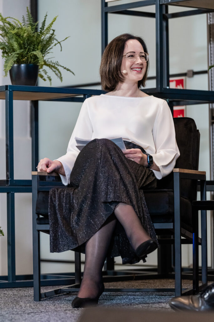 Eine Frau sitzt auf einem Stuhl in einer modernen Veranstaltungslocation. Sie trägt ein weißes Oberteil, einen schwarzen, schimmernden Rock, schwarze Strumpfhosen und schwarze Schuhe. Sie hat ein Headset-Mikrofon, hält ein Blatt Papier in der Hand und lächelt freundlich. Im Hintergrund sind grüne Pflanzen und ein Regal mit Holzelementen zu sehen, was eine professionelle, aber gemütliche Atmosphäre schafft.