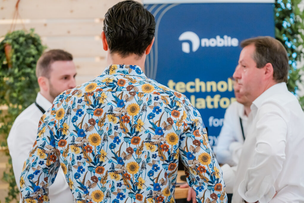 Eine Gruppe von Männern steht in angeregtem Gespräch vor einem Messestand mit dem blauen Banner der Firma 'nobile'. Im Vordergrund ist der Rücken eines Mannes in einem auffälligen, farbenfrohen Hemd mit Blumenmuster in Gelb, Blau und Orange zu sehen. Zwei weitere Männer im weißen Hemd sind im Hintergrund teilweise sichtbar. Die Szene wird von grünen Pflanzen und Holzwänden eingerahmt, was eine freundliche Atmosphäre schafft.
