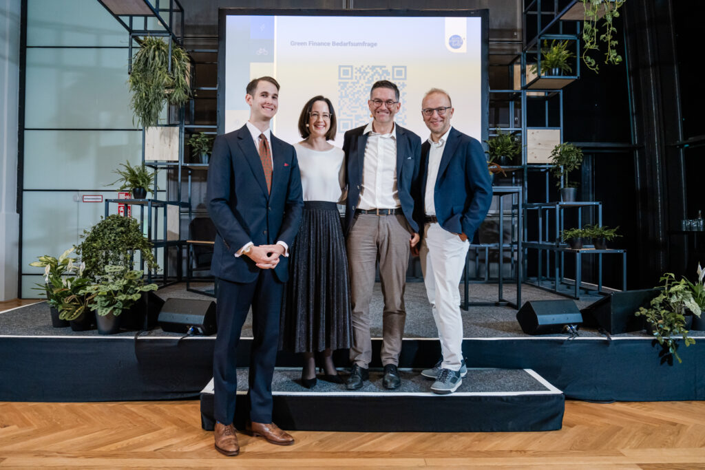 Vier Personen stehen auf einer kleinen Bühne vor einer Leinwand, auf der der Titel 'Green Finance Bedarfsumfrage' mit einem QR-Code zu sehen ist. Die Gruppe besteht aus drei Männern in Anzügen und einer Frau in einem weißen Oberteil und einem schwarzen, plissierten Rock. Sie lächeln in die Kamera und wirken entspannt. Im Hintergrund ist eine moderne, mit Pflanzen dekorierte Bühne zu sehen.