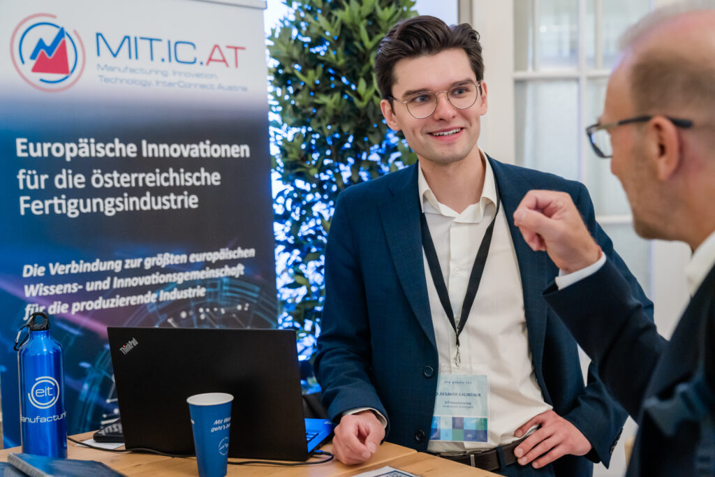Ein junger Mann im blauen Anzug, mit Brille und Namensschild, steht an einem Messestand von MIT.IC.AT, einer Initiative für europäische Innovationen in der österreichischen Fertigungsindustrie. Er lächelt freundlich und hört aufmerksam einem Gesprächspartner zu, der mit einer Geste seine Argumente unterstreicht. Auf dem Tisch vor ihnen befinden sich ein Laptop, eine blaue Trinkflasche mit der Aufschrift 'eit manufactum' und ein blauer Becher mit der Aufschrift 'Mit Wasser läuft’s besser'. Im Hintergrund ist ein Roll-up mit Informationen über das Netzwerk zu sehen.